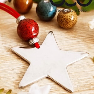 May include: A silver-coloured star-shaped pendant attached to a red cord with a red speckled bead and silver-toned spacers. The text "cdb Céline Dupré Déco" is visible in the upper right corner. The star has a smooth, metallic finish.