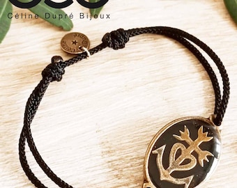 May include: Black cord bracelet with a gold-toned oval charm featuring a stylised heart, anchor, and tree design. The charm has a black enamel background. The bracelet is tied with a knot and has a small round charm with the text "Celine Dupre" on it.
