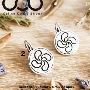 Puede incluir: Pendientes de plata con un diseño redondo que presenta una cruz vasca, también conocida como Lauburu, con un diámetro de 18 mm. Los pendientes están etiquetados como "Collection «Lauburu/ Croix basque»" y "Finition Argent".