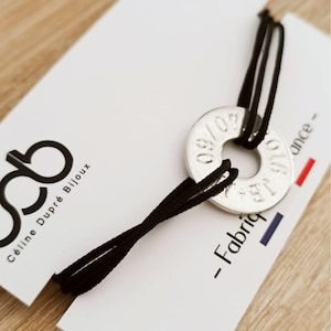 Peut inclure: Un médaillon circulaire en métal argenté gravé avec les chiffres 09/07 et les lettres JB, enfilé sur un cordon noir fin. Le médaillon est fixé sur une carte de présentation blanche portant le texte Céline Dupré Bijoux et Fabriq.