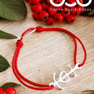 Può includere: Braccialetto in corda rossa con un ciondolo a forma di ancora in argento con un cuore. Il braccialetto è su una superficie di legno con bacche rosse sullo sfondo. Il testo "Céline Dupré Bijoux" è visibile in alto a destra.