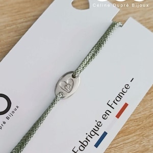 May include: A sage green cord bracelet featuring a small, oval-shaped silver-toned metal charm with an embossed heart and the number 10. The bracelet is attached to a white display card with the text Céline Dupré Bijoux and Fabriqué en France.