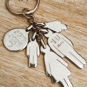 May include: A silver-toned metal keychain featuring a circular charm engraved with 2016, 2026, and a heart, alongside three stylized human figure charms. One figure is engraved with 10 Ans. The keychain includes a standard split ring attachment.