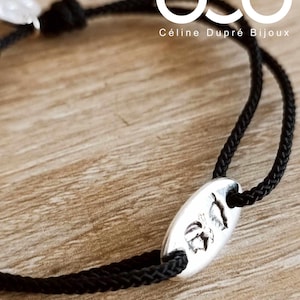 May include: Black cord bracelet with a silver oval charm featuring a detailed design. The bracelet is tied with a knot and has the text "Céline Dupré Bijoux" on the tag.