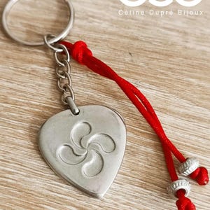 Peut inclure: Un porte-clés argenté en forme de cœur avec un symbole basque Lauburu. Il est orné d'un cordon rouge et d'un anneau métallique. Le texte "Céline Dupré Bijoux" est visible en arrière-plan.