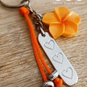 Könnte beinhalten: Ein Schlüsselanhänger mit einem silberfarbenen Metallring, einem leuchtend orangefarbenen Blumenanhänger und einem rechteckigen silberfarbenen Metallanhänger mit drei eingravierten Herzen. Eine orangefarbene Kordel mit zwei kleinen Metallperlen.
