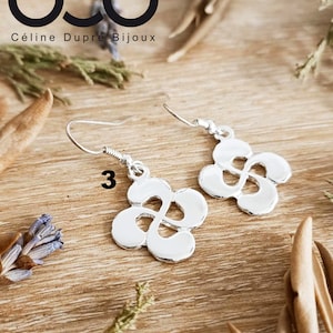 May include: Silver earrings with a delicate, intricate design. The earrings are shaped like a stylized cross, known as a Lauburu, a traditional Basque symbol. The earrings are 20 mm in diameter and have a silver finish. The earrings are part of a collection called "Lauburu/Croix Basque".