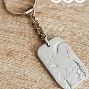 Op de afbeelding: Zilverkleurige sleutelhanger met een rechthoekige hanger met een berg- en paraglidingontwerp. De sleutelhanger bevat een splitring en een ketting. De tekst "cdb Céline Dupré Bijoux" is zichtbaar.