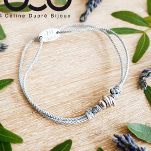 May include: A gray bracelet with a silver charm that says "10 Ans" and three silver round beads. The bracelet is on a wooden surface with green leaves and lavender flowers.
