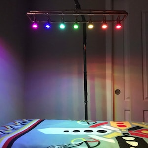 May include: A multi-colored LED light fixture with seven lights, each emitting a different color. The lights are mounted on a metal frame attached to a black pole. The lights are positioned above a bed with a patterned bedspread.