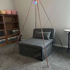 May include: A copper-colored metal pyramid structure with a multi-colored light fixture at the top. The pyramid is set up over a gray fabric chair with a pillow.