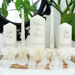 May include: A set of five cream-coloured pillar and taper candles, each adorned with a frayed-edge cream ribbon bow. The candles feature black printed text including the names Katarina and Benjamin, the date 28th February 2026, and family names.