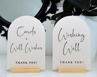 May include: Two white arch-shaped signs with black calligraphy text on wooden stands. The sign on the left says "Cards + Well Wishes Thank You!" and the sign on the right says "Wishing Well Thank You!"
