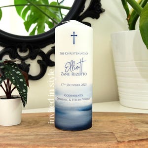May include: A white cylindrical pillar candle featuring a blue cross at the top and text that reads The Christening of Elliott Zane Rizbetto 17th October 2025 Godparents: Domenic & Helen Walker, with a blue watercolour gradient design at the base.