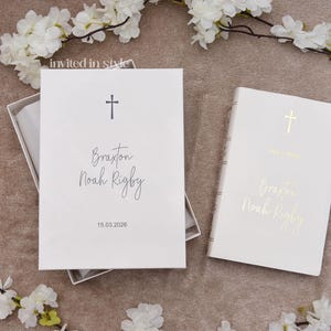 May include: A white rectangular box containing a card with a black cross, the name Braxton Noah Rigby, and the date 15.03.2026, alongside a white book featuring a gold cross and the same name in gold foil lettering, surrounded by white flowers.