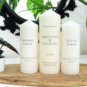 May include: Three ivory pillar candles, each with black text for a wedding. The central candle reads "The Wedding of Christine & Michael" with the date and church location. The other candles display family names. The candles are on a wooden tray.