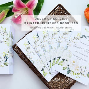 May include: White wedding booklets with floral designs, stacked and fanned out in a woven basket. The text "Order of Service" and "Printed, Finished Booklets" is visible. The booklets are decorated with white flowers and green leaves. A pink lily and other flowers are in the background.