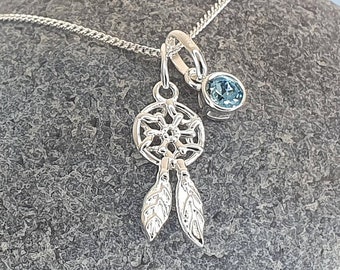 May include: A silver necklace with a dreamcatcher charm and a light blue gemstone. The necklace is on a gray rock.
