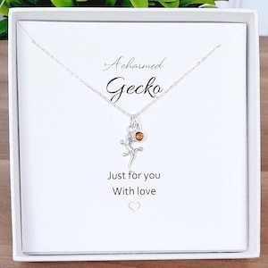 Op de afbeelding: Een witte cadeaudoos met een zilveren ketting erin. De ketting heeft een klein gecko-bedeltje met een oranje edelsteen. De doos is bedrukt met de tekst "Acharmed Gecko" en "Just for you With love".