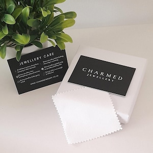 May include: A black and white card with the text "CHARMED JEWELLERY" on it. The card is sitting on a white surface with a white cleaning cloth.