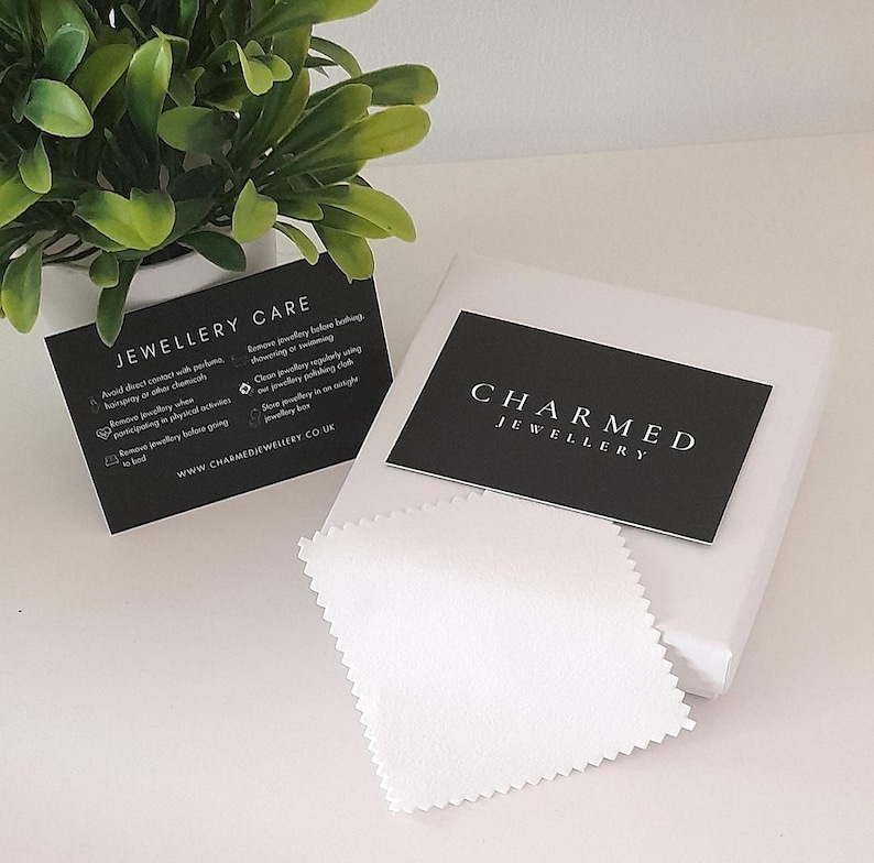 May include: A black and white card with the text "CHARMED JEWELLERY" on it. The card is sitting on a white surface with a white cleaning cloth.