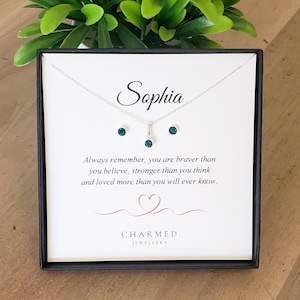 May include: A silver necklace and stud earrings set with teal gemstones, displayed in a black box. The name "Sophia" is written above a quote: "Always remember, you are braver than you believe..." The brand name "CHARMED JEWELLERY" is at the bottom.