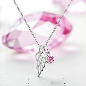 Könnte beinhalten: Eine silberne Halskette mit zarter Kette. Der Anhänger hat einen ausgeschnittenen Flügel und einen kleinen, runden, rosa Edelstein. Der Hintergrund ist in einem zarten Rosa gehalten.