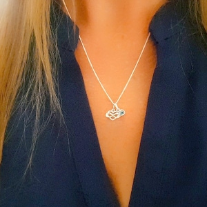 May include: A silver necklace with a small infinity symbol charm and a blue gemstone. The necklace is worn by a person with long blonde hair and a dark blue shirt.
