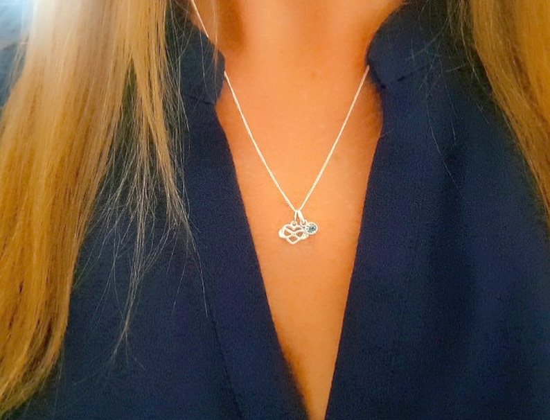 May include: A silver necklace with a small infinity symbol charm and a blue gemstone. The necklace is worn by a person with long blonde hair and a dark blue shirt.