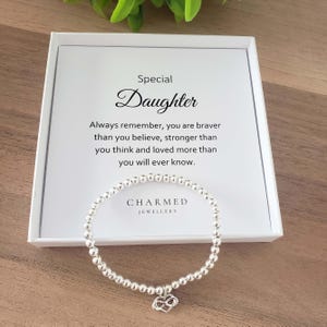Puede incluir: Una pulsera de cuentas plateadas con un dije de corazón se encuentra en una caja blanca. La caja tiene las palabras "Special Daughter" y un mensaje sentimental impreso. También se ve la marca "CHARMED JEWELLERY".