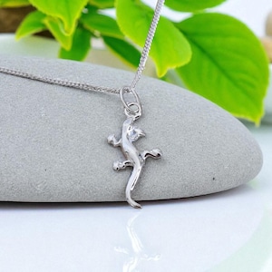 May include: A silver-toned metal necklace featuring a small, stylised lizard pendant suspended from a delicate chain. The lizard charm has a smooth, reflective surface and is attached to the chain by a simple circular jump ring connector.