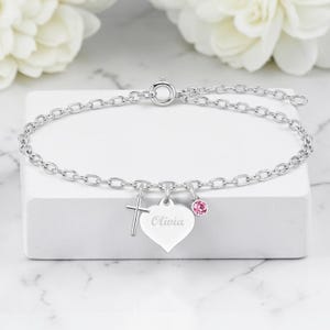 May include: A silver bracelet with a cross charm, a heart charm engraved with "Olivia", and a pink gemstone charm. The bracelet is displayed on a white surface with white flowers in the background.