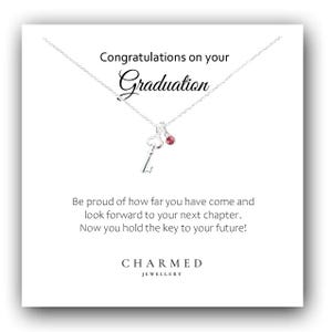 Op de afbeelding: Een zilveren ketting met een sleutelhanger en een kleine roze edelsteen, gepresenteerd op een witte kaart. De kaart leest "Gefeliciteerd met je afstuderen" en bevat inspirerende tekst. De merknaam "CHARMED JEWELLERY" staat onderaan.