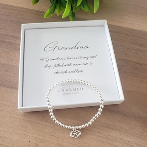 Pode incluir: Uma pulseira de contas prateadas com um berloque de coração está em frente a uma caixa branca. A caixa tem a palavra "Avó" em escrita cursiva, com uma mensagem sobre amor e memórias. A marca "CHARMED JEWELLERY" também é visível.