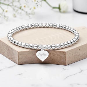 Könnte beinhalten: Ein silberfarbenes Perlenarmband mit polierter Oberfläche und einem herzförmigen Anhänger. Der Anhänger ist mit dem Namen Sophia in Schreibschrift graviert. Das Armband besteht aus einer durchgehenden Reihe kleiner, gleichmäßiger, runder Perlen.