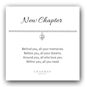 Könnte beinhalten: Ein silbernes Armband mit einem Herzanhänger, präsentiert auf einer weißen Karte. Die Karte zeigt den Schriftzug "New Chapter" in Schreibschrift, zusammen mit inspirierendem Text und dem Logo von "Charmed Jewellery".