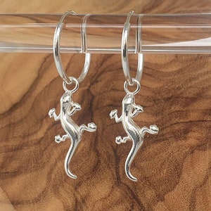 May include: A pair of silver hoop earrings with a silver lizard charm hanging from each hoop.