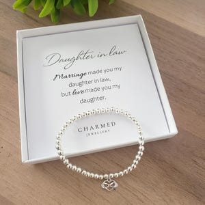 Puede incluir: Una pulsera de cuentas plateadas con un dije en forma de corazón, presentada en una caja de regalo blanca. La caja tiene el texto "Daughter in law" y un mensaje. La pulsera es de Charmed Jewellery.