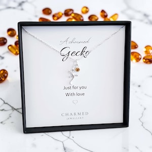 May include: A silver-toned necklace featuring a small gecko pendant and a single amber-coloured crystal charm. The jewellery is presented in a black box with a white card printed with the text A charmed Gecko, Just for you, With love, and CHARMED JEWELLERY.