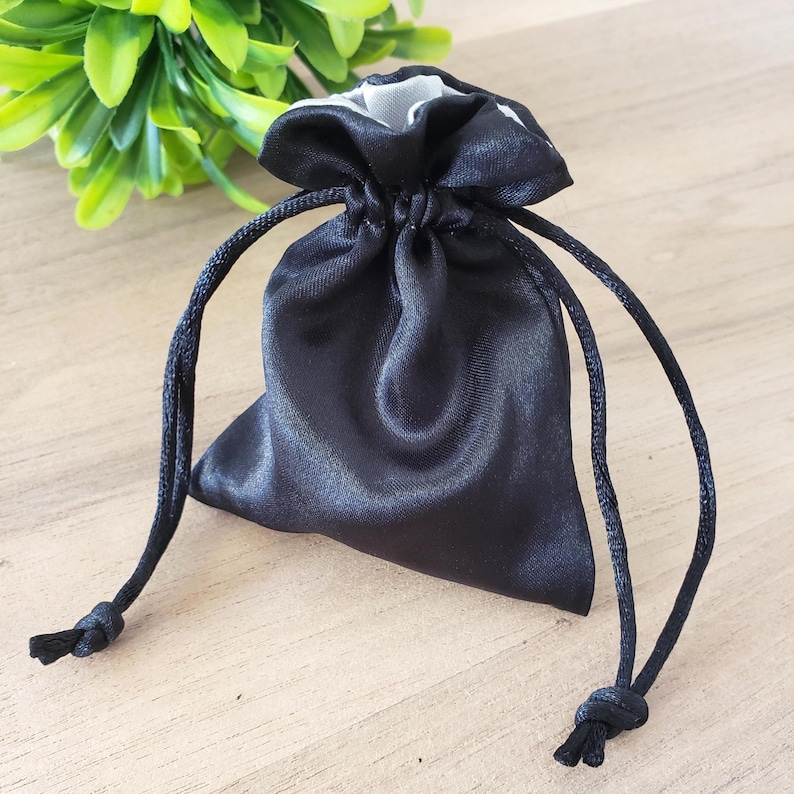 May include: A small, black satin drawstring bag with a contrasting white interior. The bag is gathered at the top with a black cord, tied with knots at the ends. The bag is on a light-coloured wooden surface.
