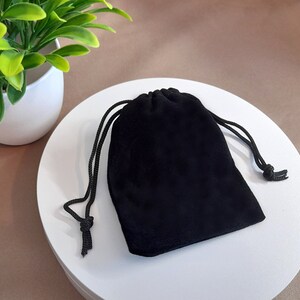 May include: A black velvet drawstring bag, closed with a black cord. The bag is sitting on a white surface, with a small potted plant in the background.