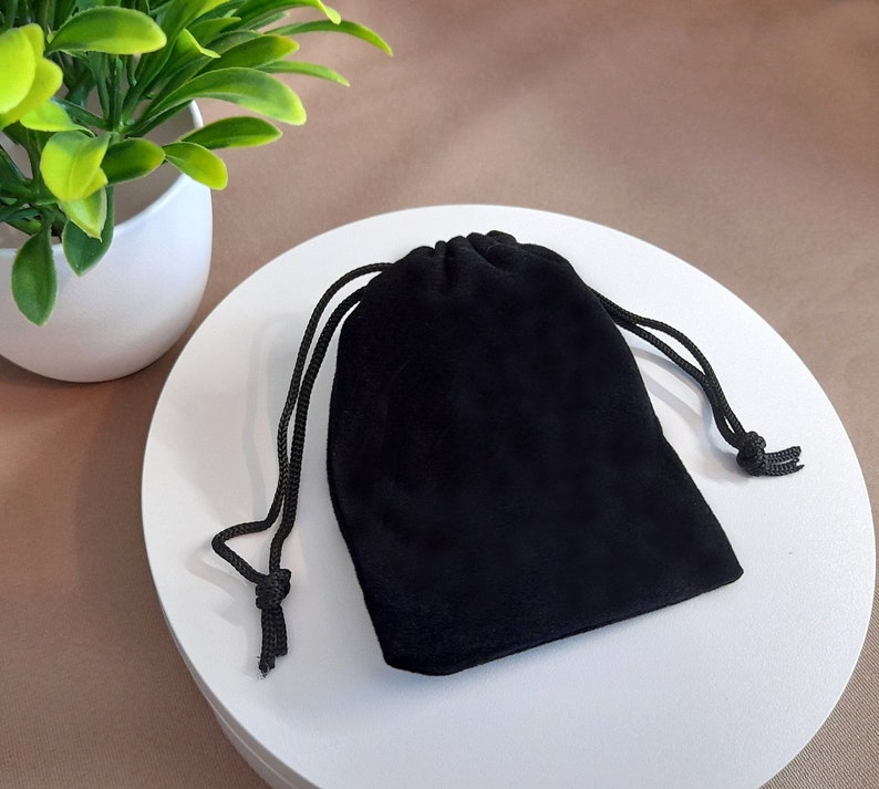 May include: A black velvet drawstring bag, closed with a black cord. The bag is sitting on a white surface, with a small potted plant in the background.