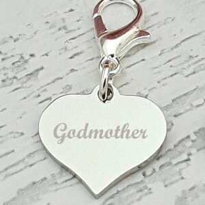 May include: A silver heart-shaped charm with the word "Godmother" engraved on it. The charm is attached to a silver clasp. The background is a light gray and white wood grain pattern.