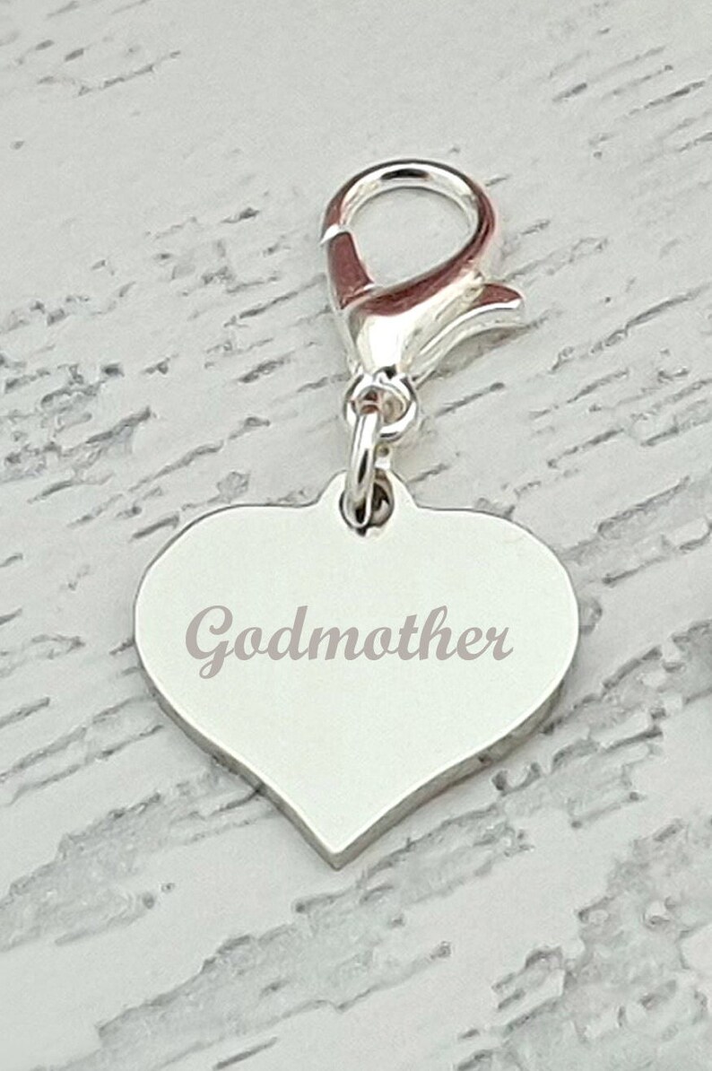 May include: A silver heart-shaped charm with the word "Godmother" engraved on it. The charm is attached to a silver clasp. The background is a light gray and white wood grain pattern.
