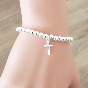 Puede incluir: Pulsera plateada con cuentas redondas y un dije de cruz. La pulsera presenta pequeñas cuentas redondas y un sencillo colgante de cruz, creando un accesorio religioso delicado.