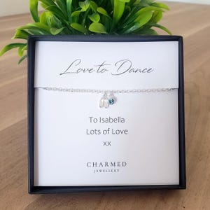 Puede incluir: Una pulsera de plata con dos colgantes, una zapatilla de ballet y una gema azul, se muestra en una caja negra. El texto "Love to Dance" está encima de la pulsera, con "To Isabella Lots of Love" debajo. La marca "CHARMED JEWELLERY" está en la parte inferior.