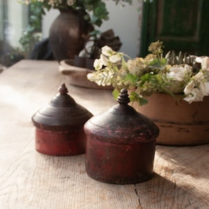 May include: Two small, round, wooden boxes with lids. The boxes are painted a deep red colour and have a distressed finish. The boxes are sitting on a wooden table.