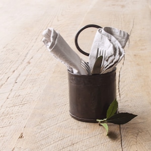 May include: A black metal utensil holder with a ring on top. A white linen napkin is tucked into the holder, along with a fork and knife. A green leaf is in the foreground.