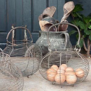 May include: A vintage wire egg basket containing brown eggs, with a metal handle. Several other wire baskets are in the foreground. A large, dark grey ceramic jug holds wooden spoons. The scene is set on a wooden surface.