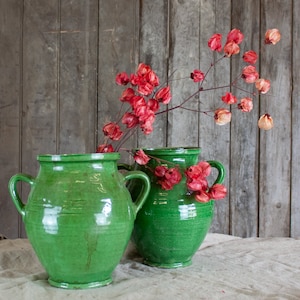 May include: Two green ceramic vases with handles, containing branches of red dried flowers. The vases are a bright, glossy green and sit on a neutral cloth. The background is a weathered wooden wall.
